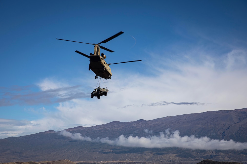 A vehicle attached to a helicopter flies over mountains against a cloudy, blue sky.