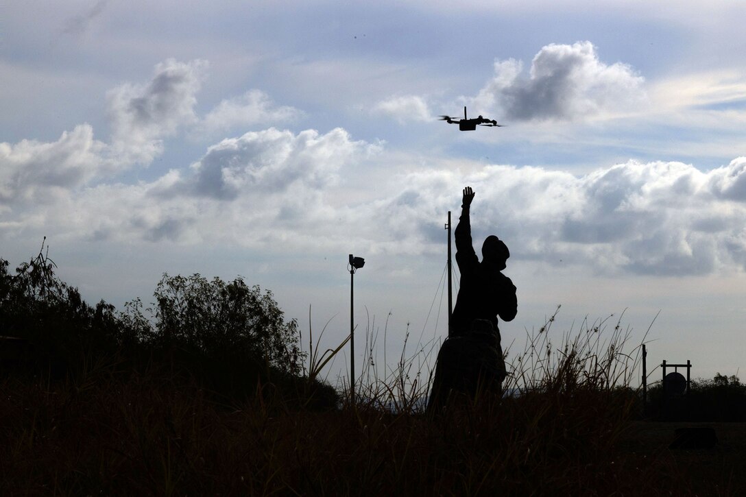 A Marine in silhouette stands around low shrubs and reaches up with one arm to release a small drone against cloudy skies.