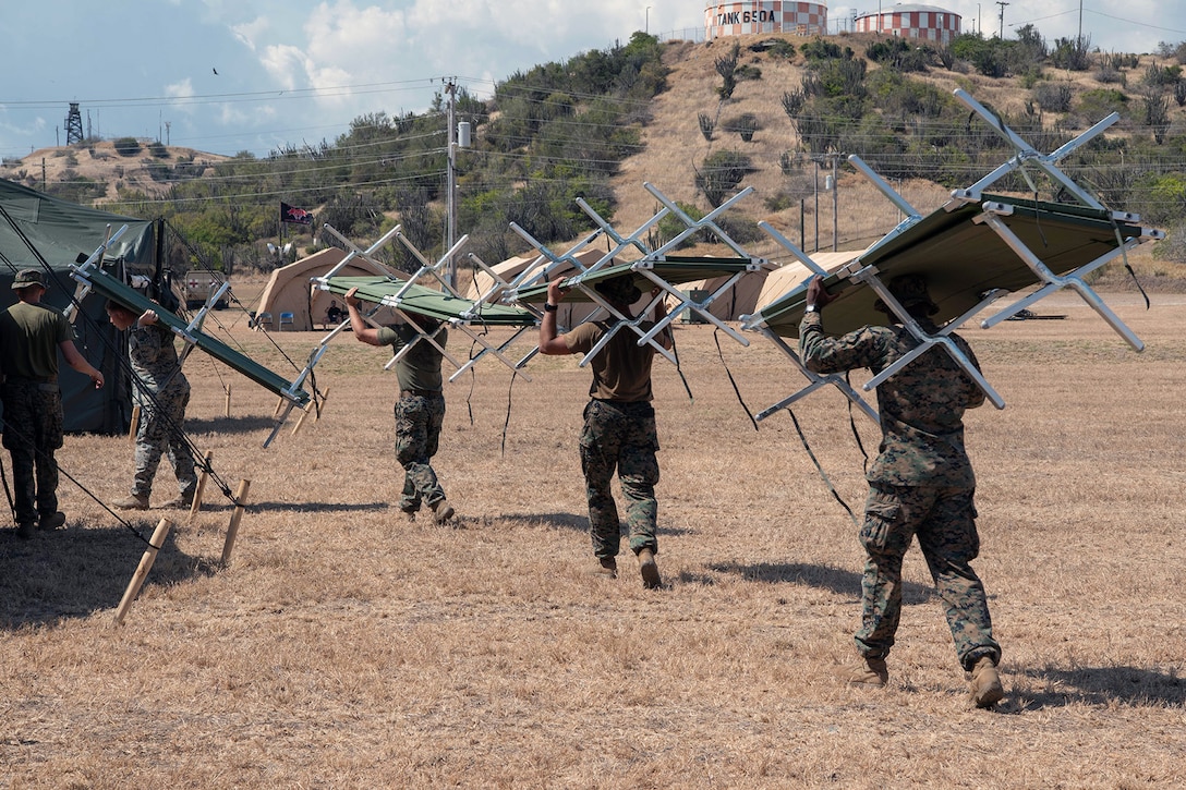 A line of Marines carry tent equipment on their heads and shoulders in an open field with hills in the backdrop.