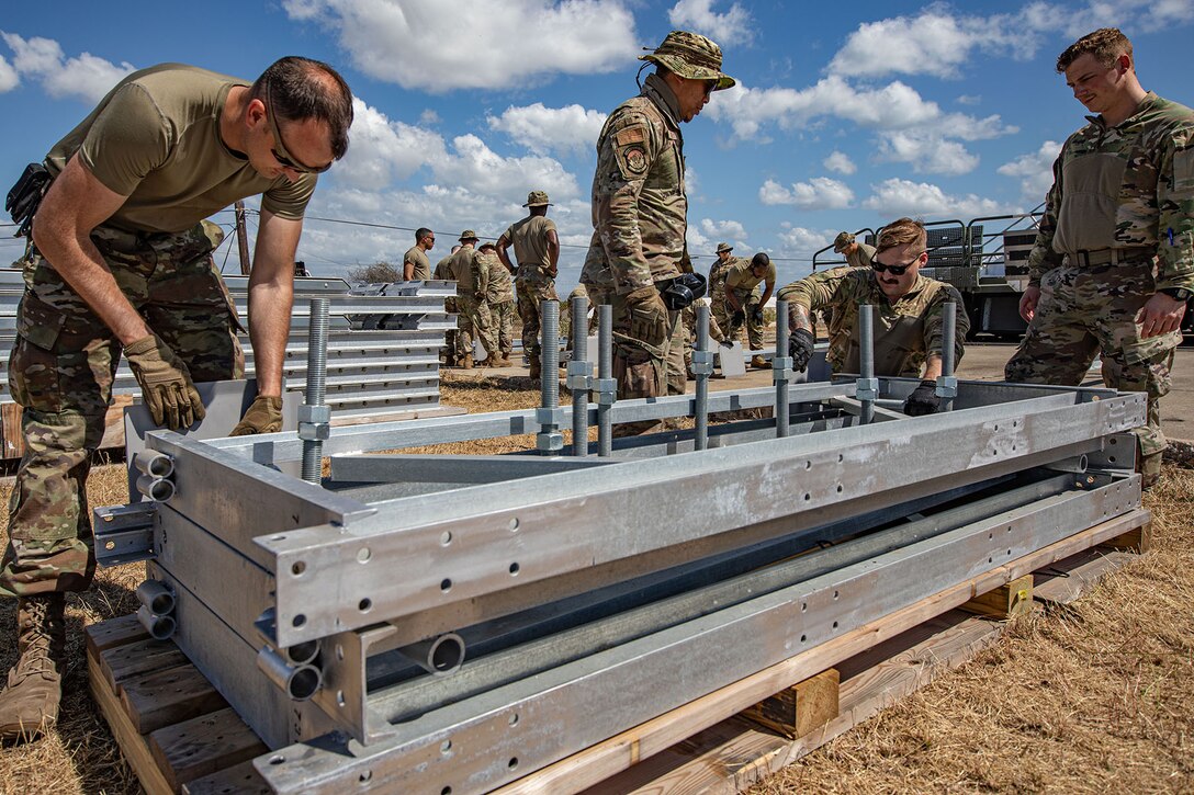 Airmen work around several metal docks atop wooden pallets in a wide area against a partly cloudy sky.