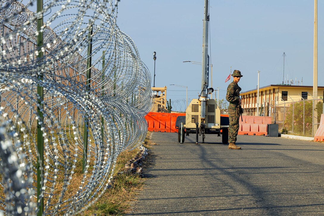 A Marine stands in the road at an installation entrance with a wire fence  off to the side.