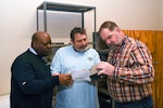 Three men stand and look a a sheet of paper with some printed text on it and a small box with a label on it.