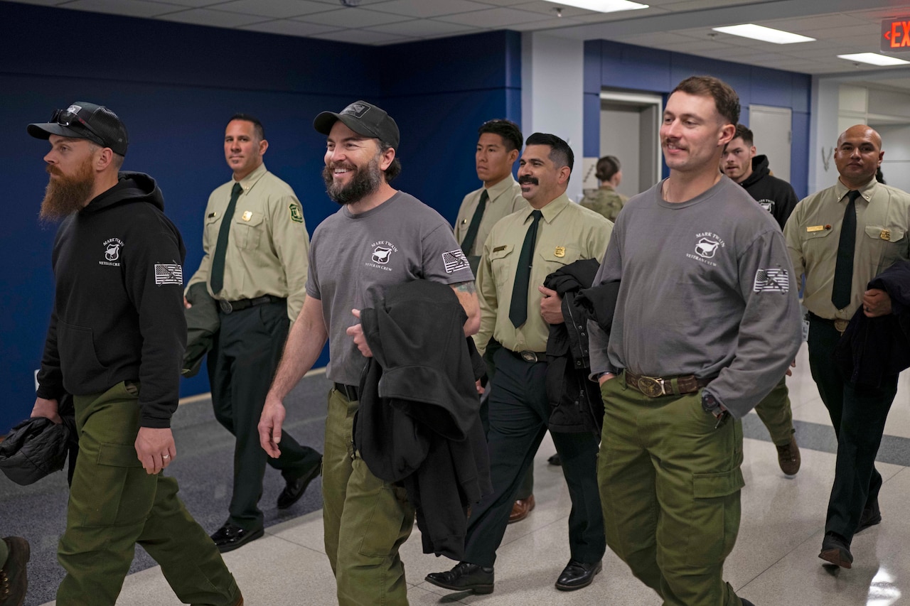 A group of men, some in dress firefighter uniforms and others in long sleeve shirts and cargo pants, walk down a hallway.