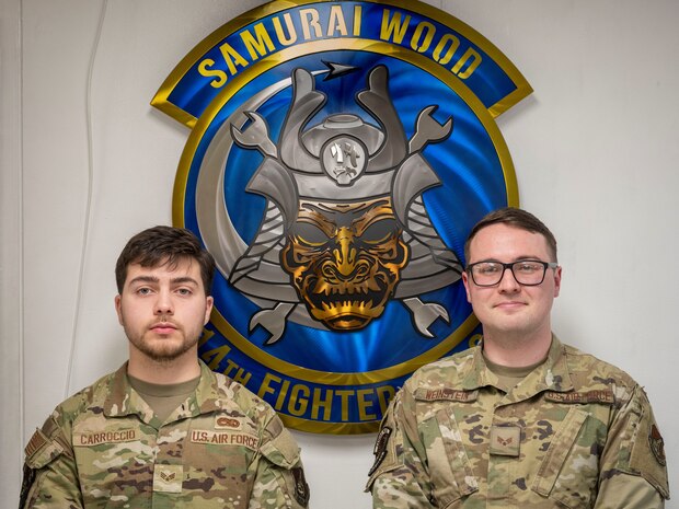 U.S. Air Force Senior Airman Chris Carroccio, left, and Senior Airman Max Weinstein, 14th Fighter Generation Squadron (FGS) aerospace propulsion technicians, pose in front of the 14th FGS logo.
