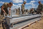 Airmen work around several metal docks atop wooden pallets in a wide area against a partly cloudy sky.
