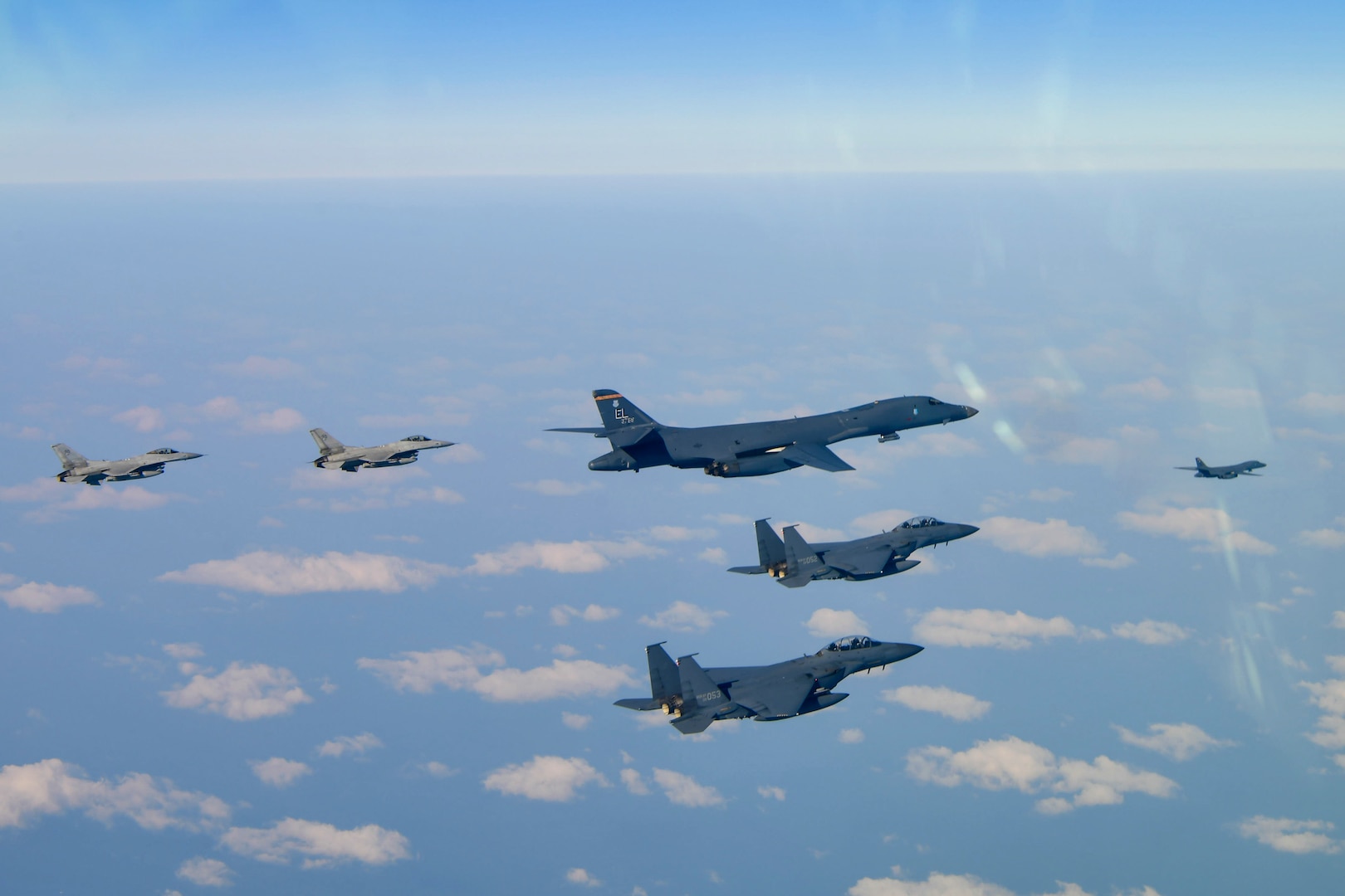 One bomber and four fighter aircraft fly in a v-formation, with another bomber out front, over scattered clouds and ground, with the horizon line at the top of the photo.