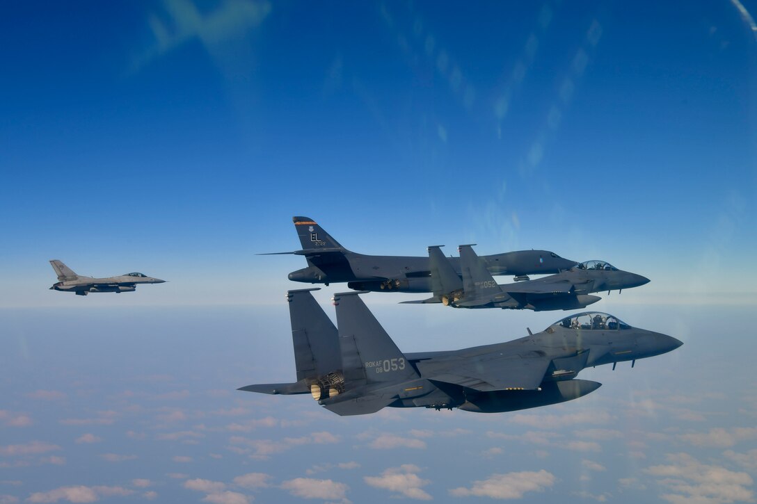 Three fighter jets and a B-1 bomber fly in the gray-blue sky with scattered clouds and the ground below.