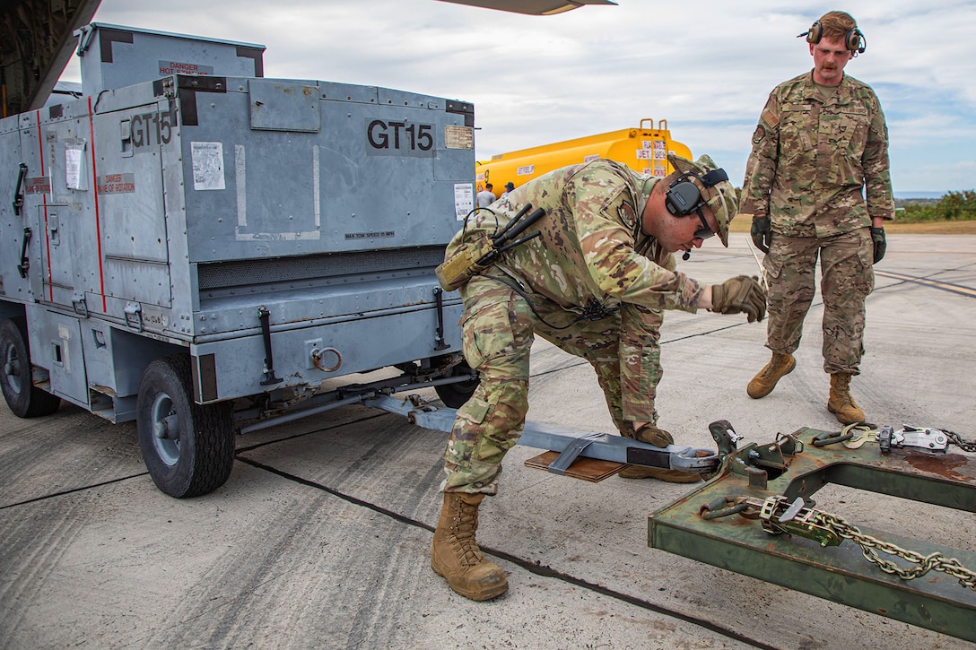 Two airmen stand between a military vehicle and a trailer hitch under cloudy skies.