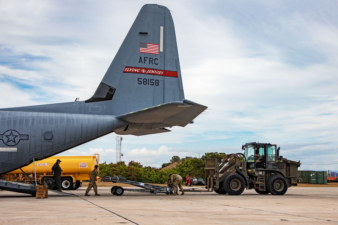 Three airmen move equipment from an open aircraft to a vehicle on a runway under partly cloudy skies.