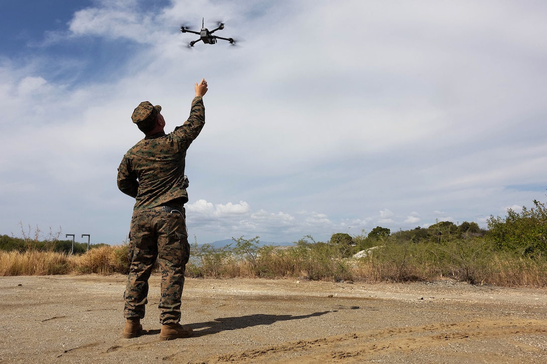 A Marine stands on asphalt in front of brush on a partly cloudy day and reaches up towards a drone.