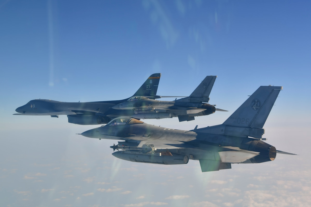 Two gray fighter jets and a B-1 bomber are shown from the side, in close formation, with a blue sky in the upper background and scatter clouds under the aircraft.