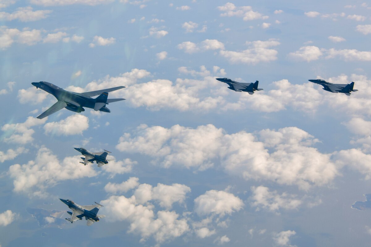One B-1 bomber aircraft, with wings forward, flies in a v-formation with two F-16s on the left and two F-15Ks on the right, over scattered, puffy clouds and a mix of water and small islands.