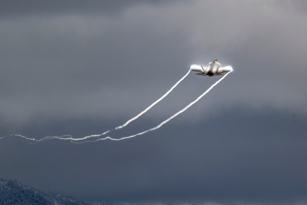 A military aircraft ascends into a dark and cloudy sky, leaving streaks of white in its wake.