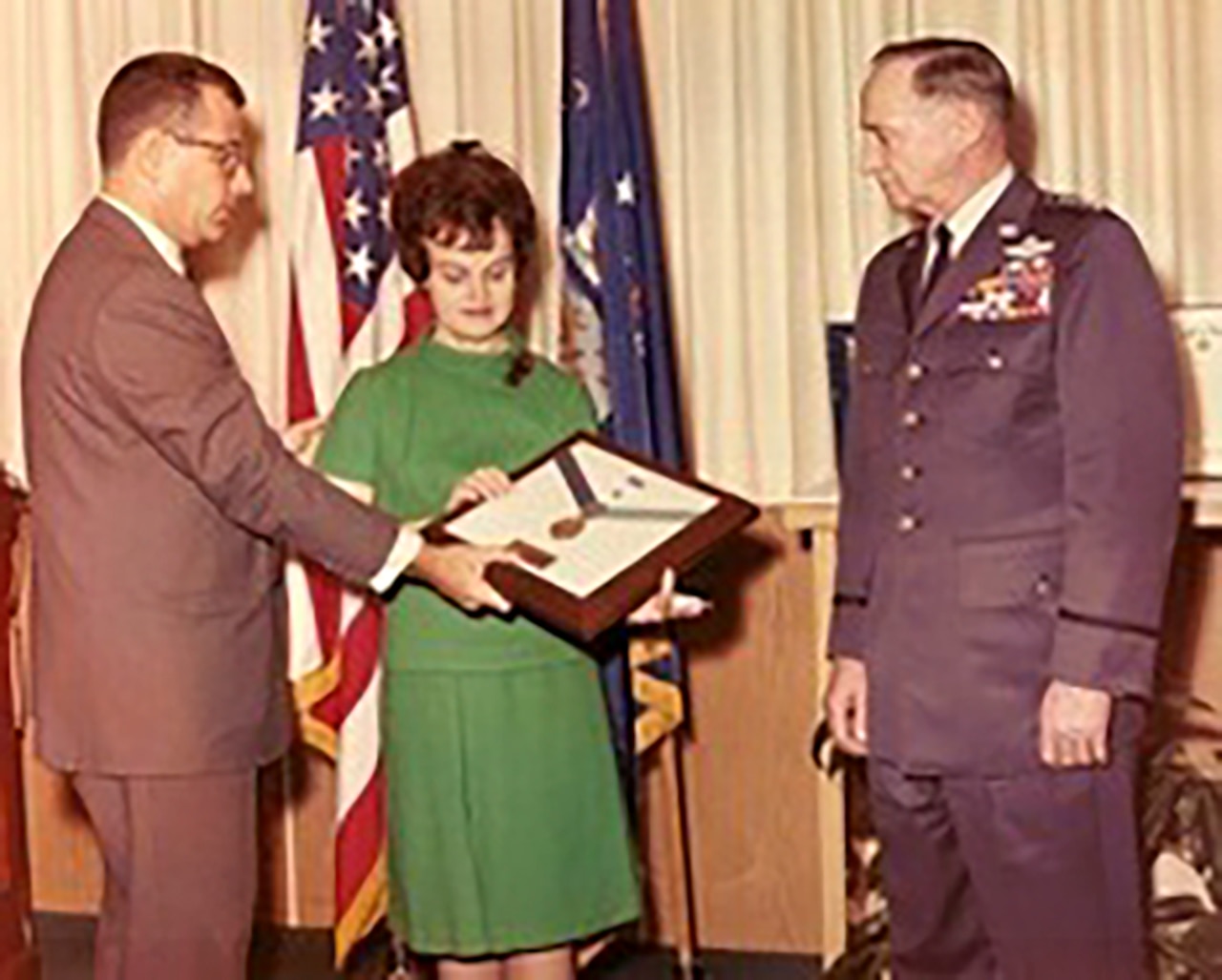 A person hands a shadow box with a medal inside it to another person as third person watches nearby.