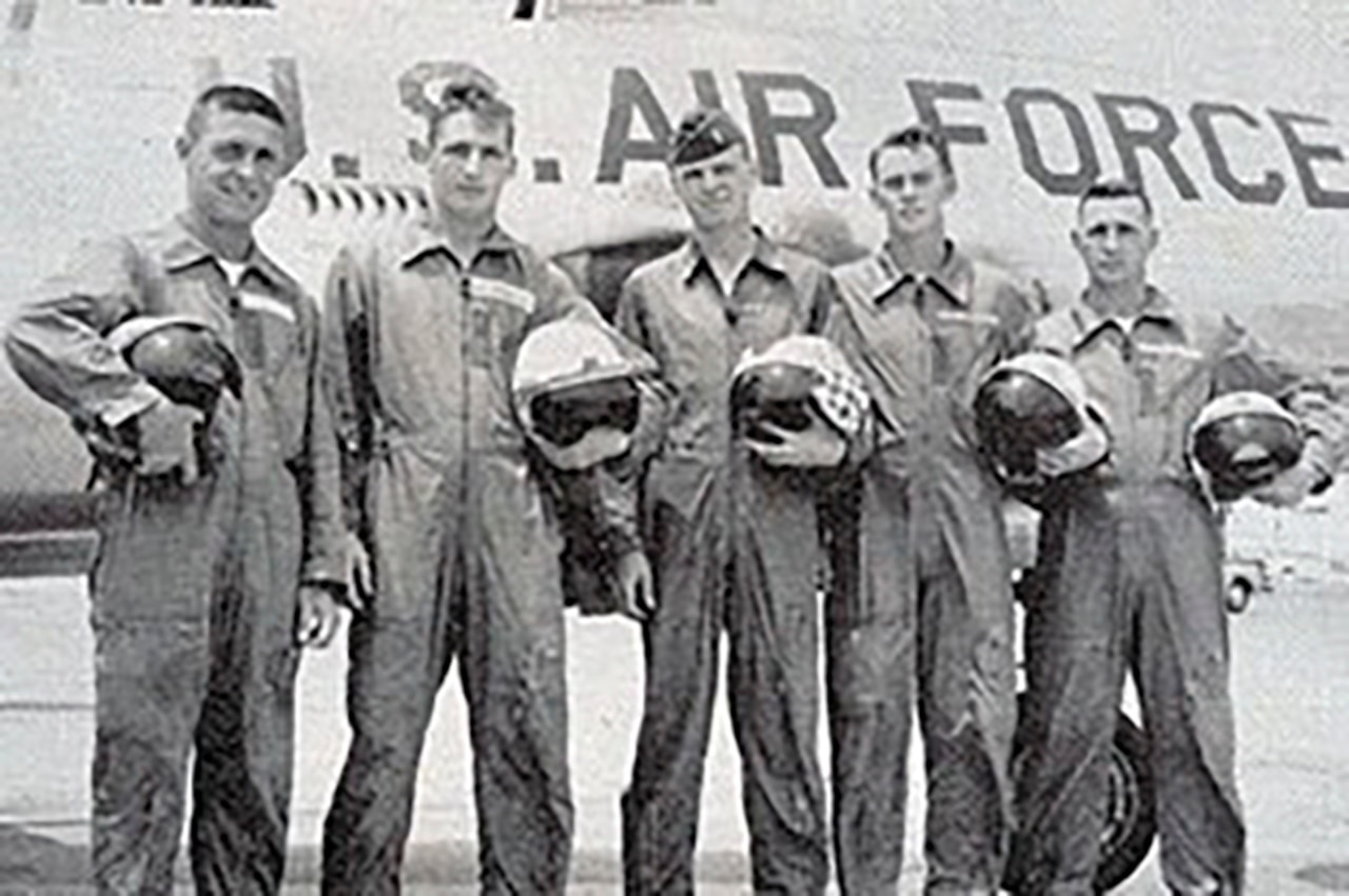 Five men in jumpsuits who are holding helmets pose in front of an aircraft.