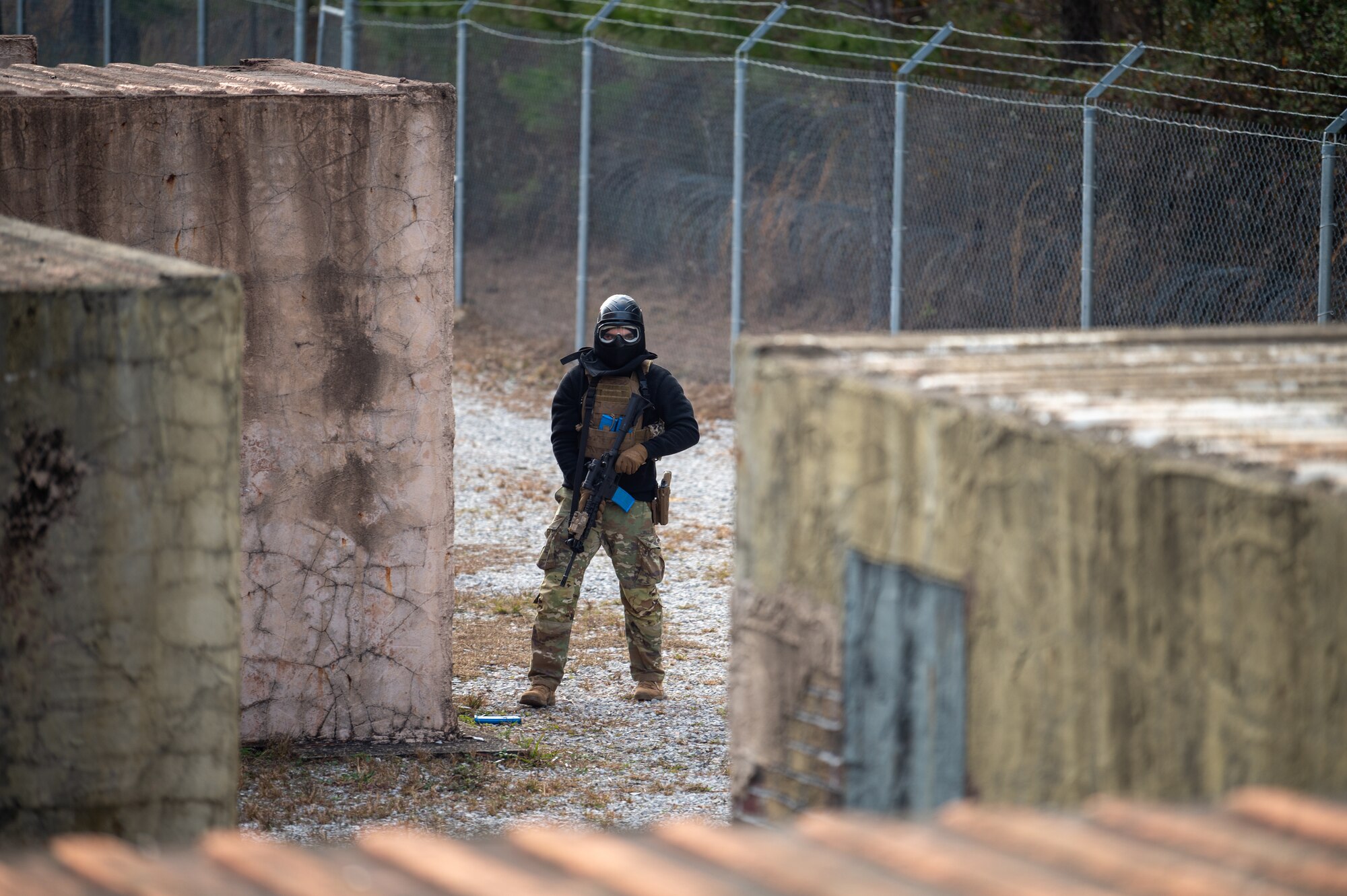 An Airmen acting as an opposing force during an FTX is in between buildings observing his surroundings while holding a rifle.