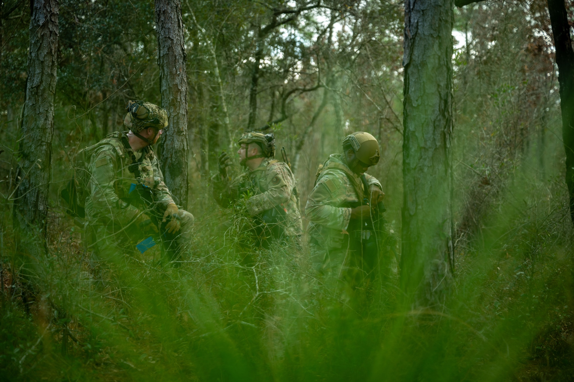 Three Airmen in the woods in a kneeling position keeping a look out. One of the Airmen is giving the hand signal for others to don their protective masks.