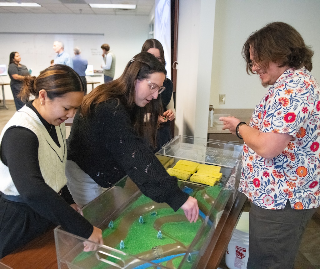 two women place small houses inside a terrain model while a man describes the model to them