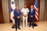 A main in a white navy uniform is flanked by 2 men in blue suits standing in front of the flags of Panama and the United States.