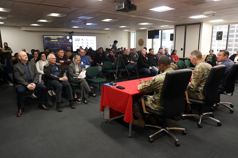 Three men sit a at a table facing a room of people sitting in chairs during a meeting.