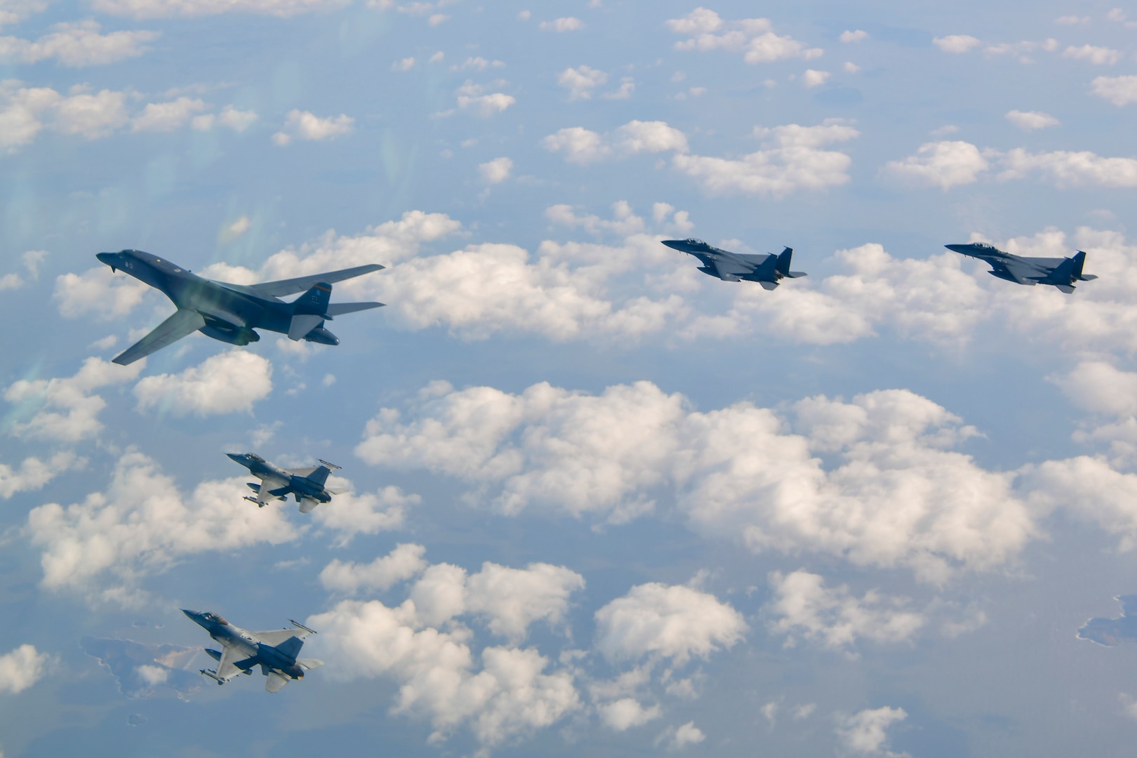 One B-1 bomber aircraft, with wings forward, flies in a v-formation with two F-16s on the left and two F-15Ks on the right, over scattered, puffy clouds and a mix of water and small islands.