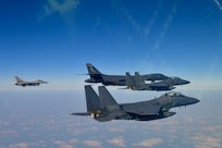 Three fighter jets and a B-1 bomber fly in the gray-blue sky with scattered clouds and the ground below.