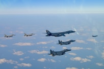 One bomber and four fighter aircraft fly in a v-formation, with another bomber out front, over scattered clouds and ground, with the horizon line at the top of the photo.