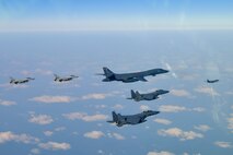 One bomber and four fighter aircraft fly in a v-formation, with another bomber out front, over scattered clouds and ground, with the horizon line at the top of the photo.