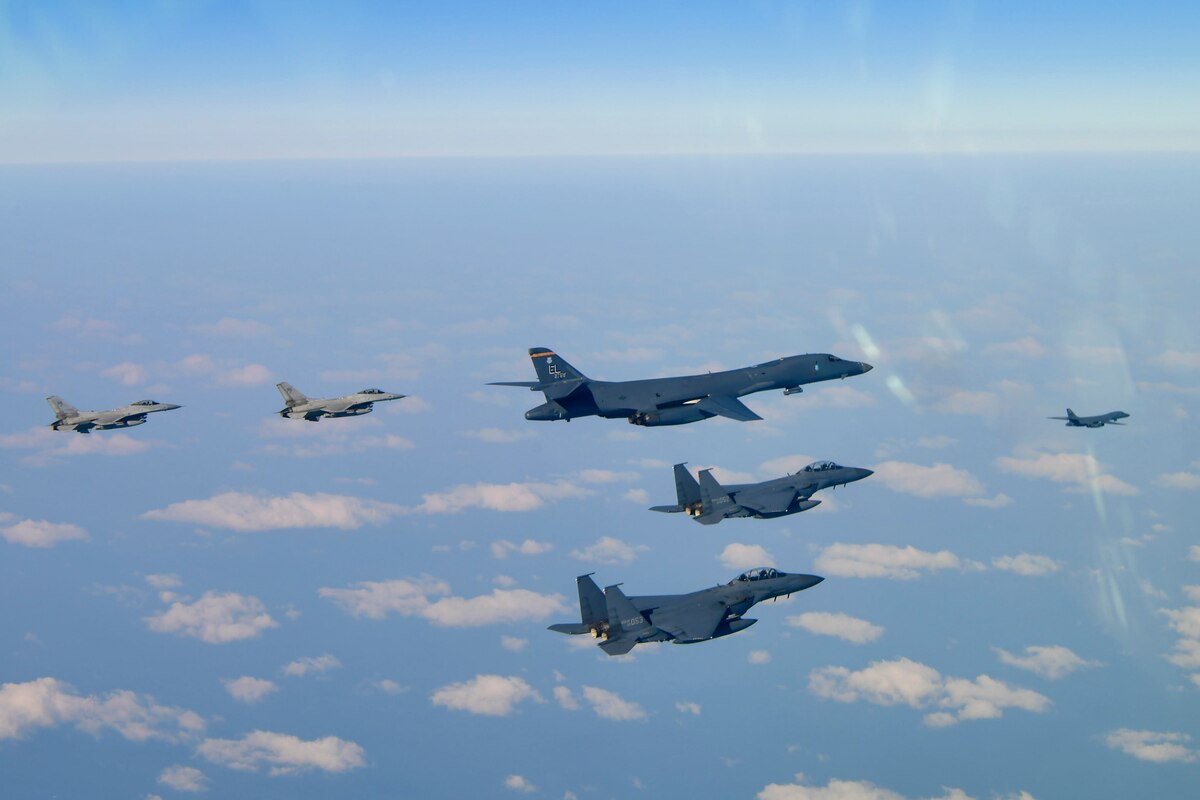One bomber and four fighter aircraft fly in a v-formation, with another bomber out front, over scattered clouds and ground, with the horizon line at the top of the photo.