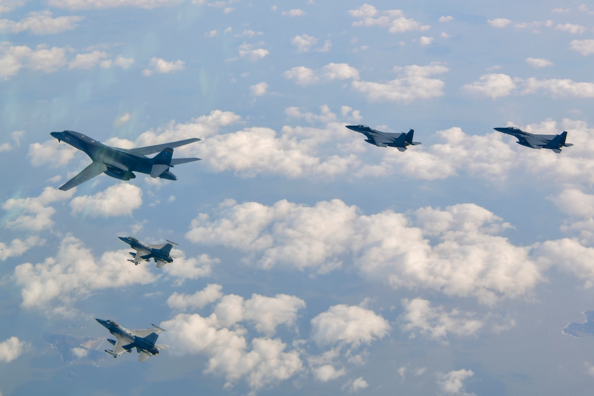 One B-1 bomber aircraft, with wings forward, flies in a v-formation with two F-16s on the left and two F-15Ks on the right, over scattered, puffy clouds and a mix of water and small islands.
