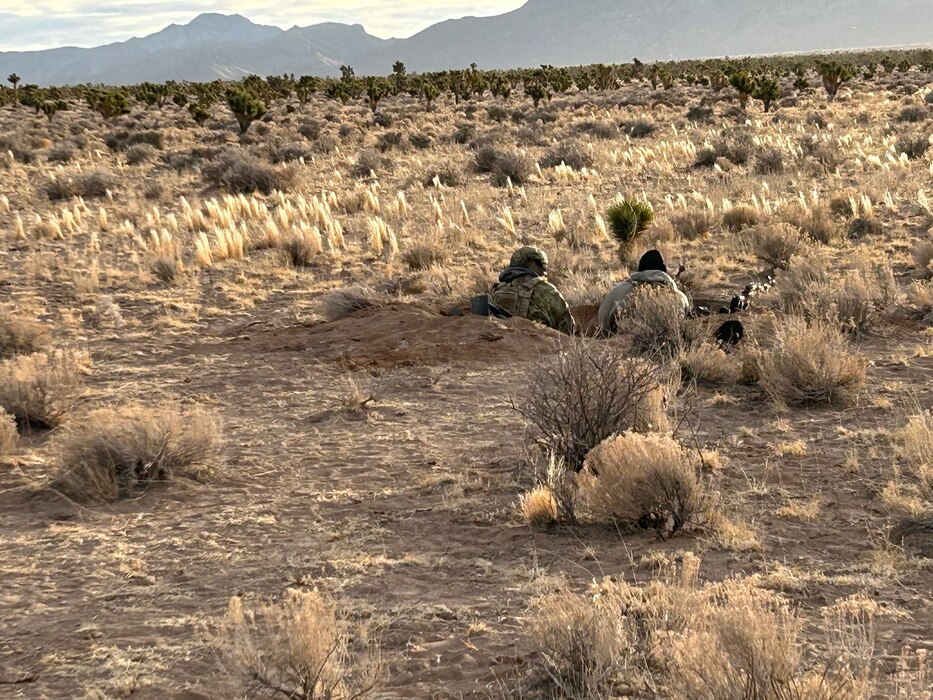 two uniformed military members crouched in a hole in a field