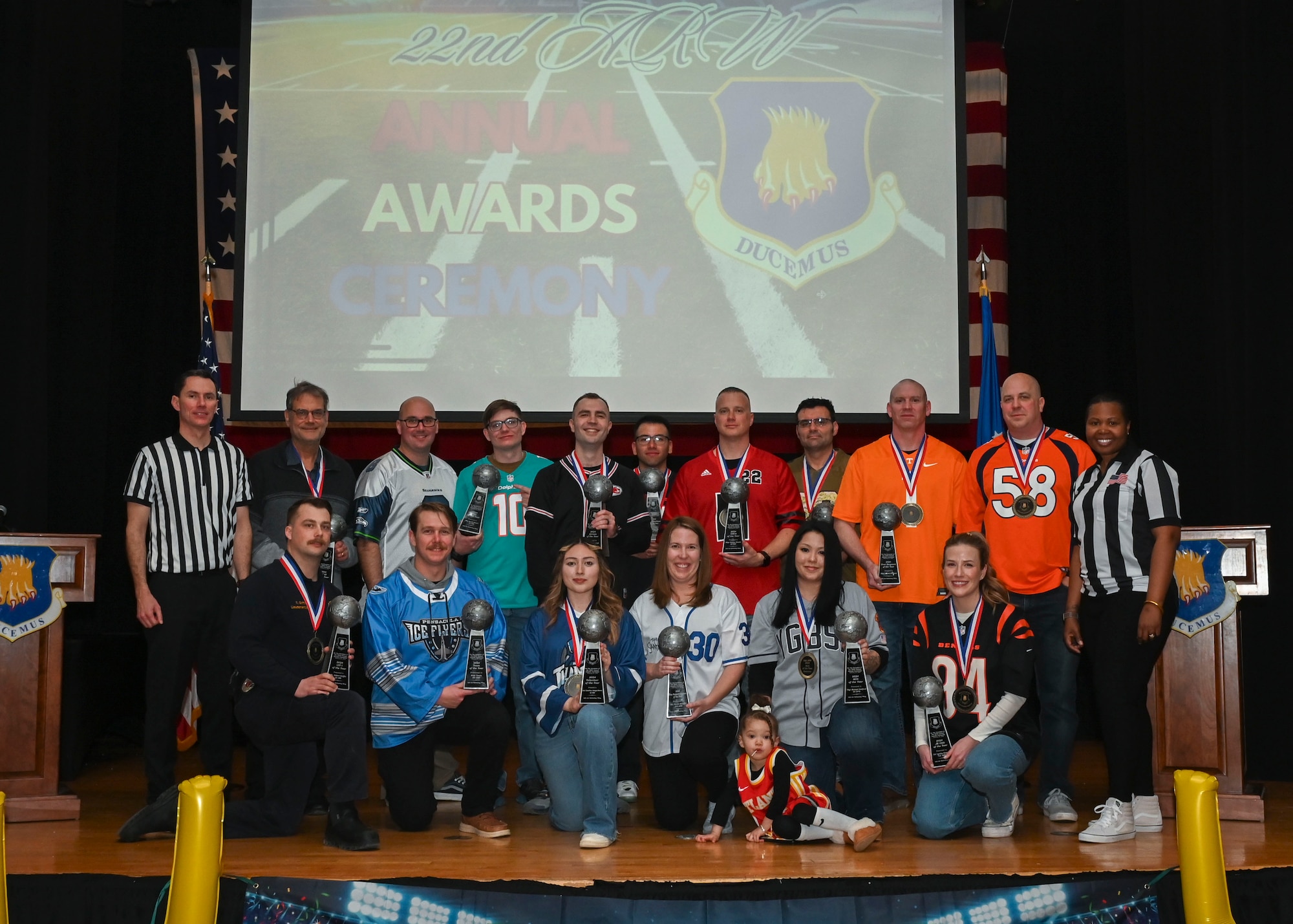 A group of people in sports jerseys pose for a group photo while holding trophies.