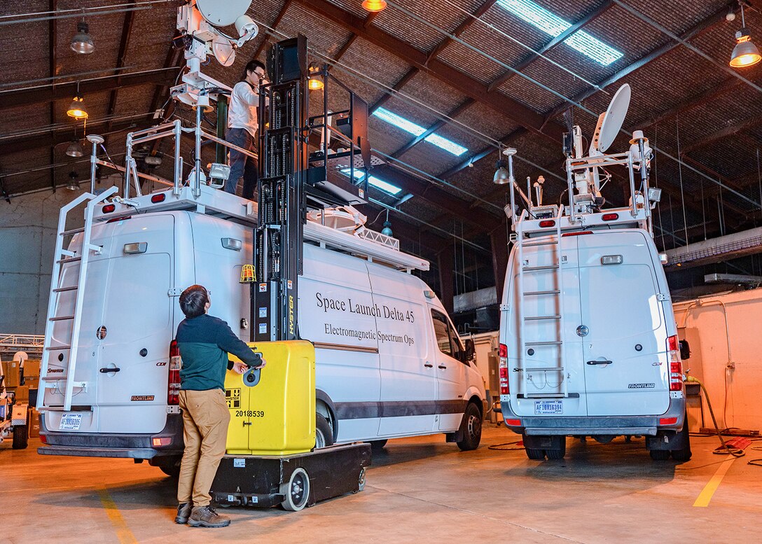 Two men work next to a van with a manlift. One of the men is on top of the van's roof.