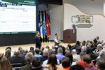 Man in suit and woman in military uniform stand in front of stage speaking to auditorium full of seated people.