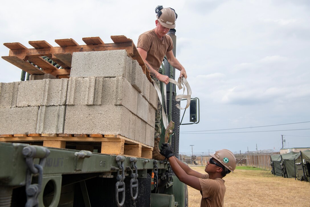 A service member stands on the bed of a truck while another works on equipment from the ground.