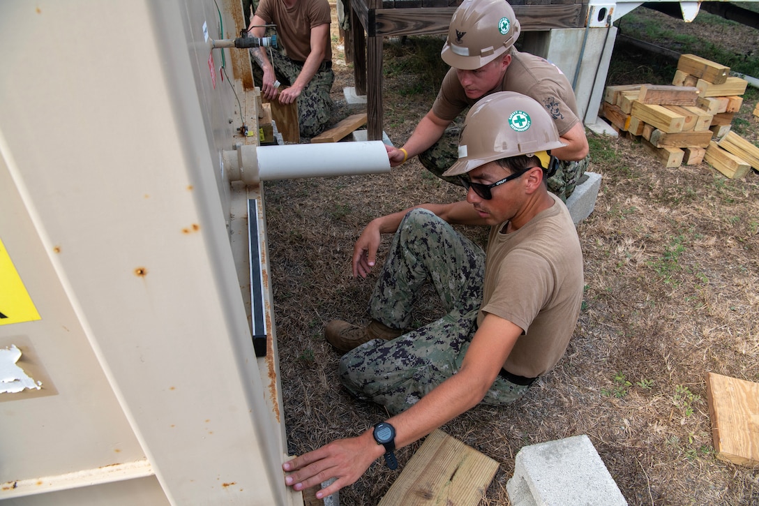 Two soldiers crouch and work on installing equipment on the side of a metal building.