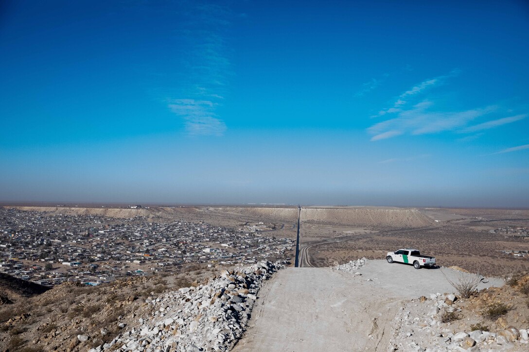A truck sits on a hill overlooking a wide arid valley with a town of small buildings in the distance and a big blue sky.