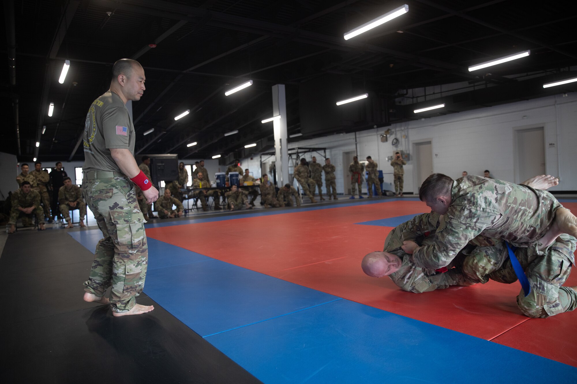Two service members grapple on floor mats while another service members observes on the far left.