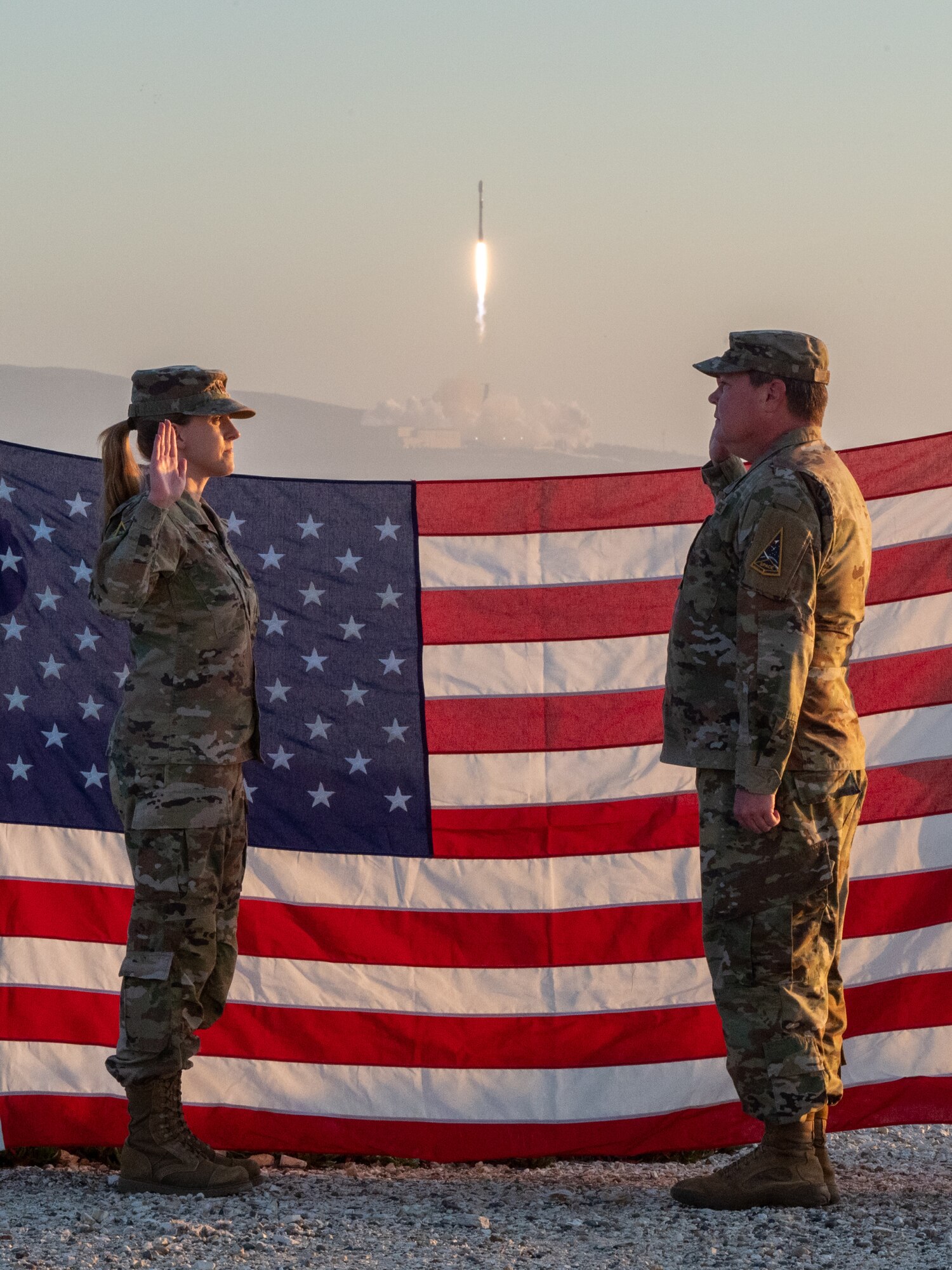 Two people stand in front of a flag while a rocket launches in the background.