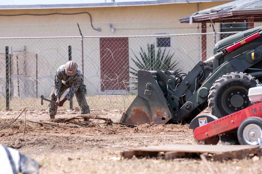 A sailor hacks at the ground while a tractor excavates near by.