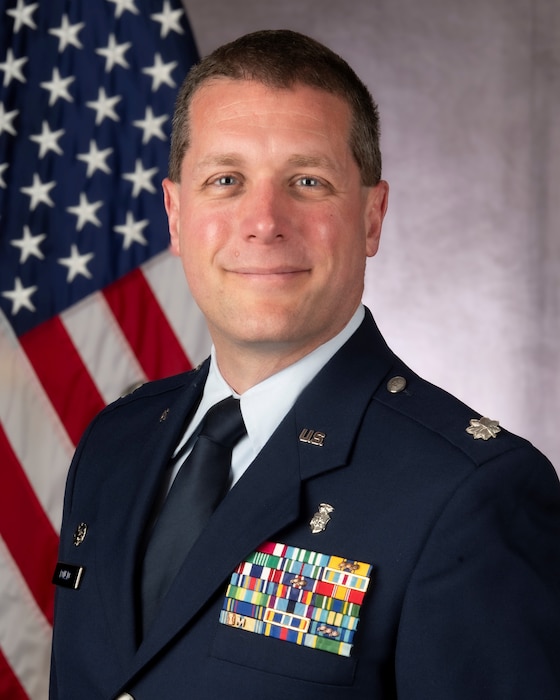 a head-and-shoulders photo of man wearing a USAF uniform in front of a flag and gray background
