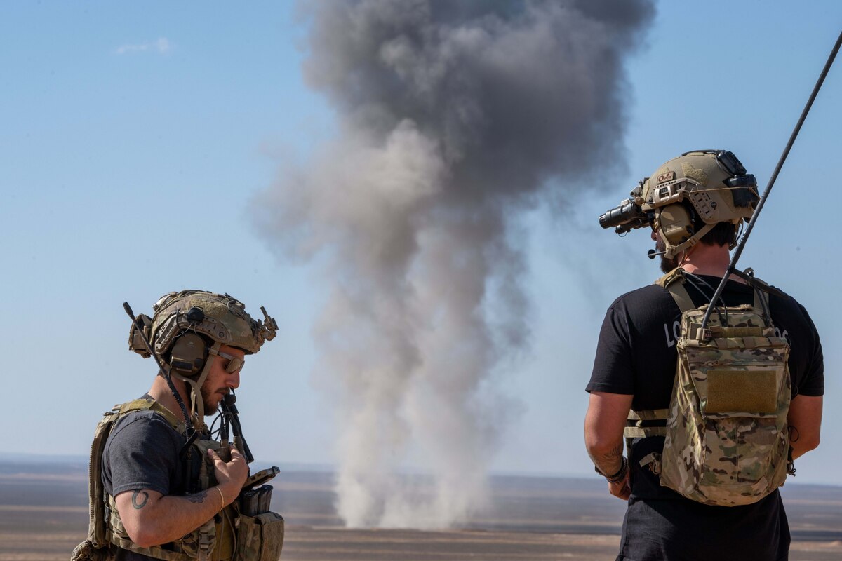 Two airmen with radio equipment stand and monitor a flat, empty field as smoke billows from the ground.