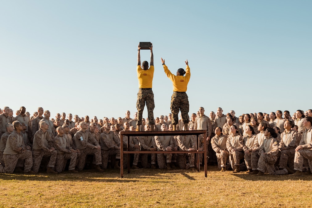 Two drill instructors wearing yellow shirts stand on a wooden platform surrounded by Marines in gear. One instructor holds a box aloft.