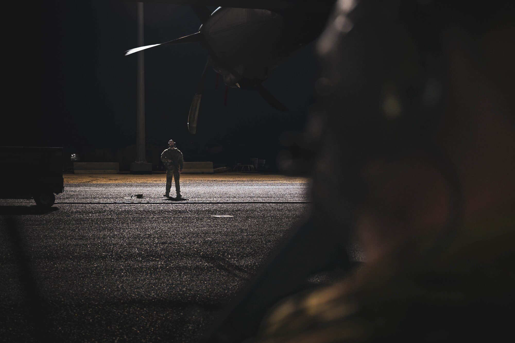 A man in uniform stands under a floodlight on a flightline while holding a rifle.