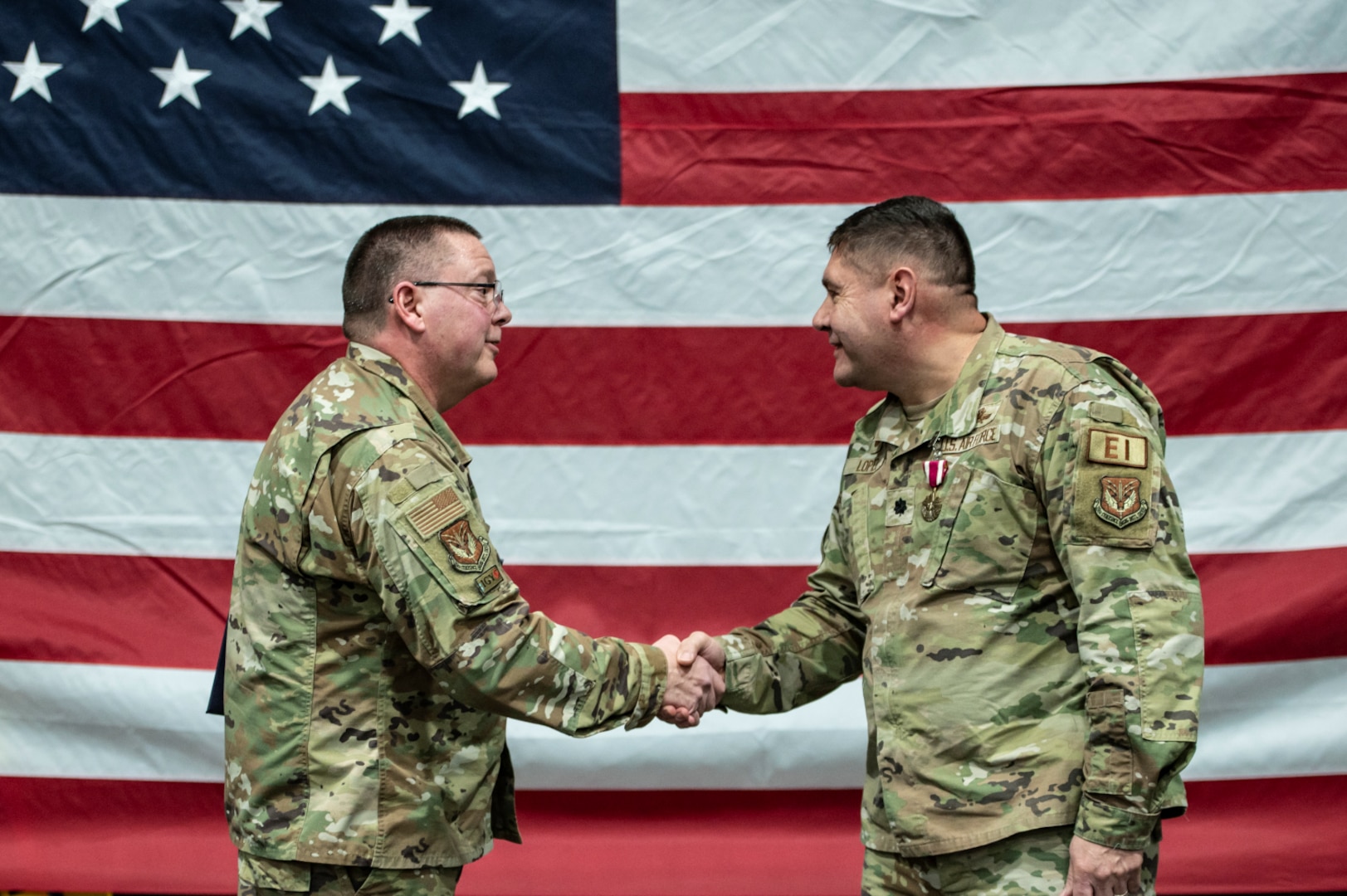 Two uniformed service members shake hands in front of the United States flag.