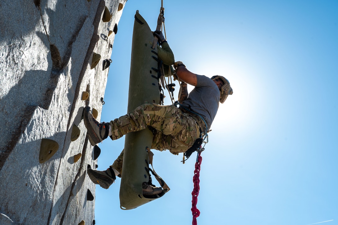 An airman in a harness attached to a fellow service member in a carrier climbs a rock wall as the sun shines in a blue sky.