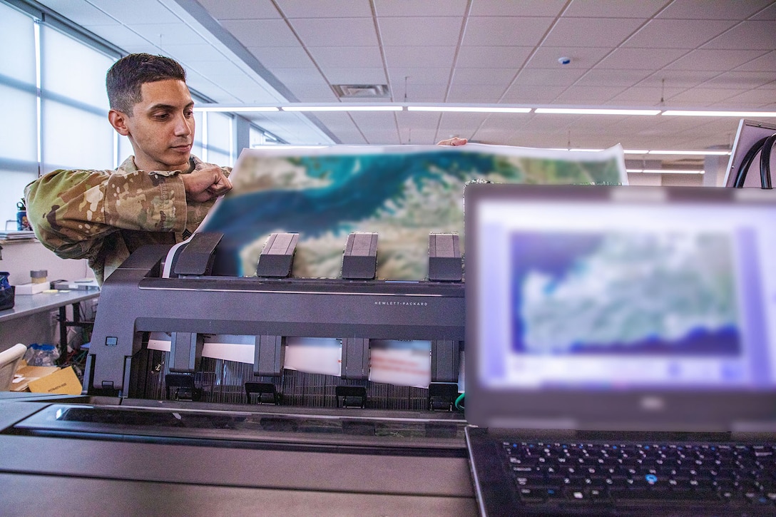 A geospatial engineer takes a map off a map printer while indoors at an office.