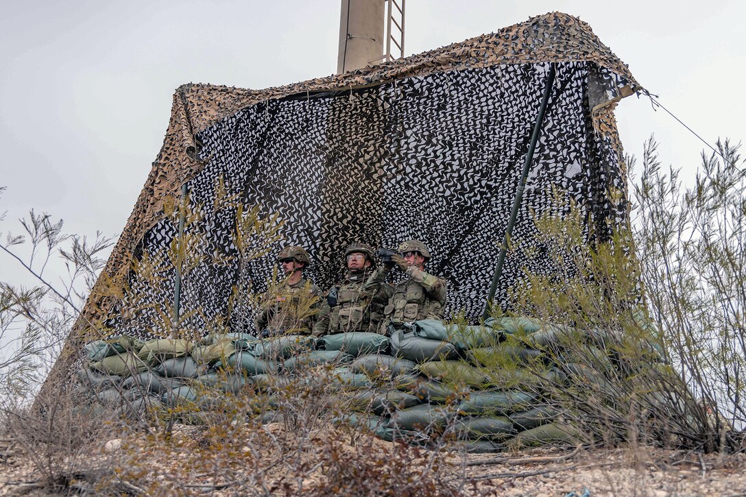Three soldiers stand behind sandbags in a makeshift enclosure and observe.