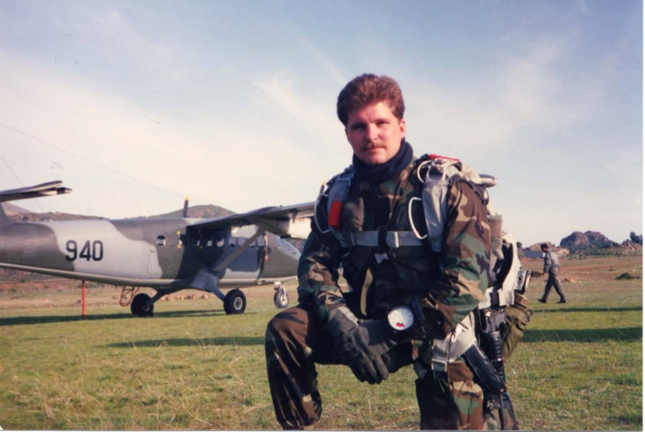 A man in parachute gear kneels on one knee near a propeller aircraft.