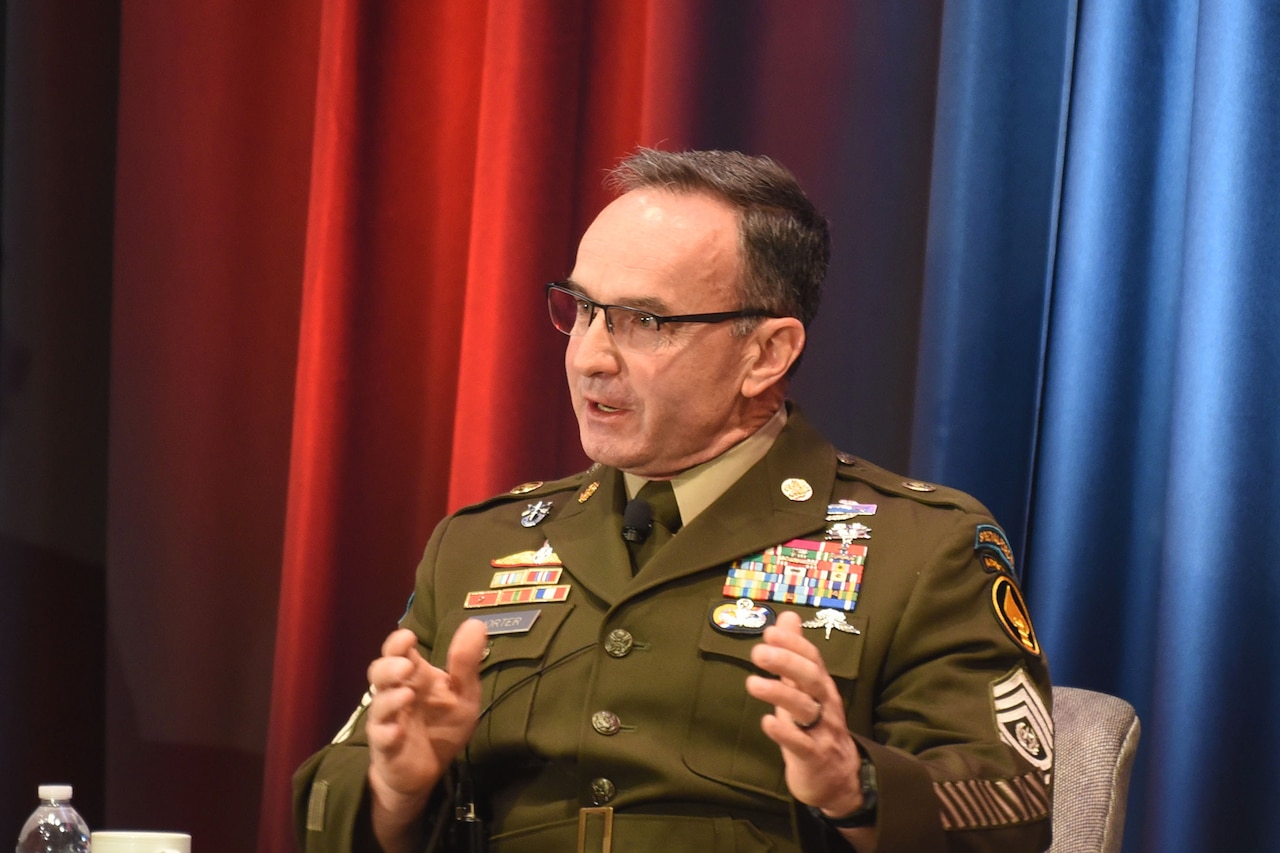 A soldier sits  in front of red and blue curtains and speaks to an audience, not pictured.